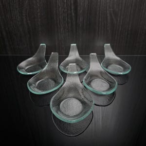 Könnte beinhalten: Sechs klare Servierlöffel aus Glas mit tropfenförmiger Form und hellgrünem Rand. Die Löffel sind auf einer schwarzen Oberfläche angeordnet, die das Licht reflektiert. Ideal zum Servieren von Vorspeisen.