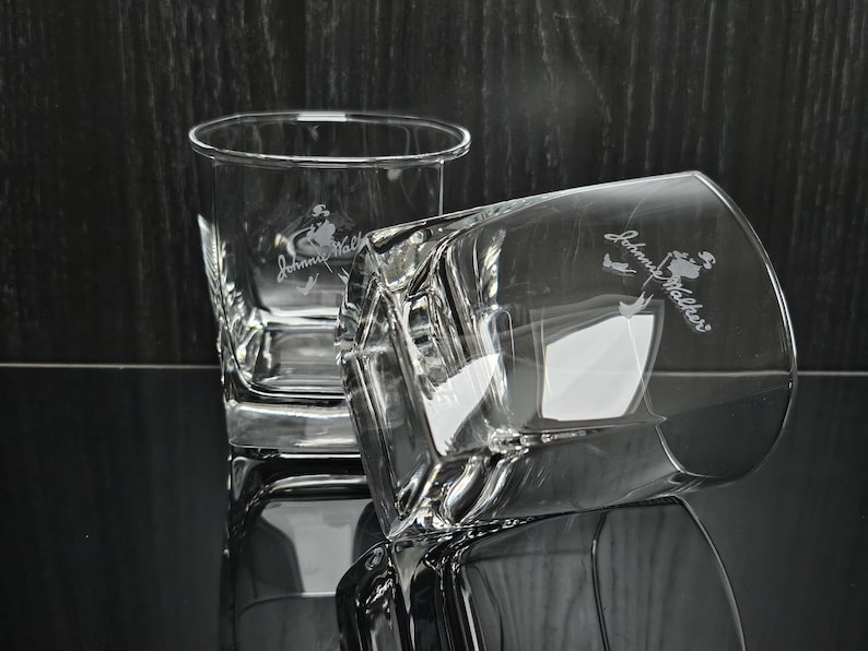 May include: Two clear, square-shaped whiskey glasses with the "Johnny Walker" logo etched on the side. One glass is upright, the other is tilted, reflecting light. The glasses are on a reflective surface.