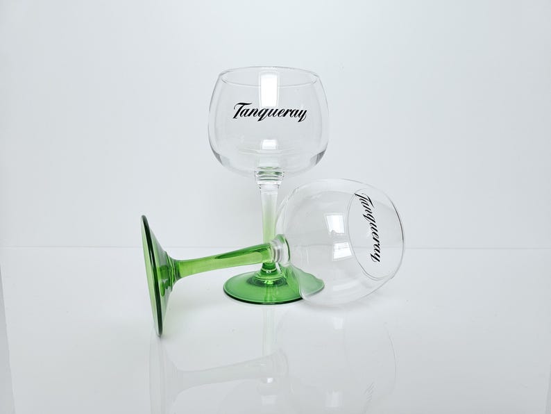 May include: Two clear, stemmed gin glasses with green bases and stems, one upright and one on its side. The word "Tanqueray" is printed in black on the glass. The glasses are on a white surface.