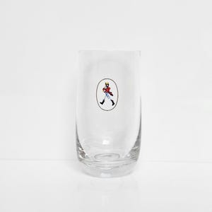 May include: A clear, cylindrical glass with a rounded base. The glass features a small, oval-shaped emblem with a cartoon figure in a red coat, black boots, and a yellow hat. The figure is walking. The glass is empty.