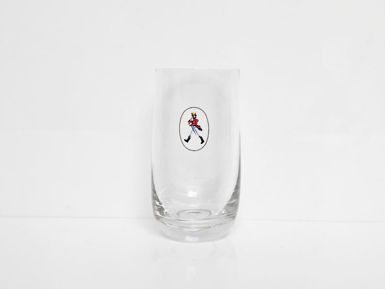 May include: A clear, cylindrical glass with a rounded base. The glass features a small, oval-shaped emblem with a cartoon figure in a red coat, black boots, and a yellow hat. The figure is walking. The glass is empty.