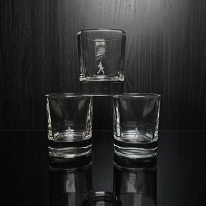 May include: Three clear, square-shaped shot glasses stacked on a reflective surface. Each glass has a small, etched logo of a figure walking. The glasses are empty, and the background is a dark wood.