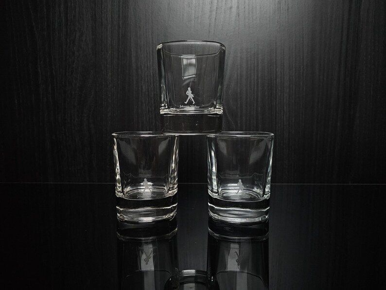 May include: Three clear, square-shaped shot glasses stacked on a reflective surface. Each glass has a small, etched logo of a figure walking. The glasses are empty, and the background is a dark wood.