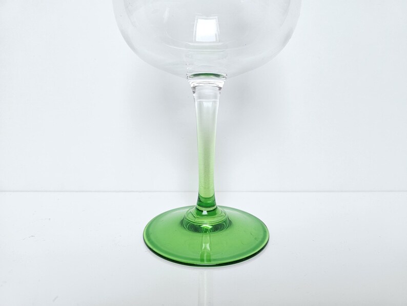 May include: A clear glass goblet with a green stem and base. The stem transitions from clear to green. The goblet has a large, rounded bowl, ideal for serving wine or cocktails. The base is a solid, circular shape.