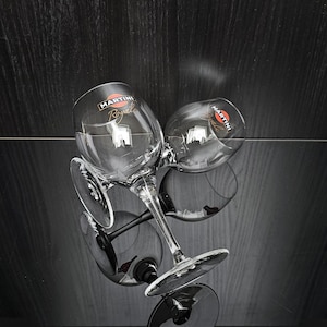May include: Two clear Martini Royal stemmed glasses with the Martini logo, on a reflective black surface. The glasses are angled, showing their design and the reflection. The background is a dark wood grain.