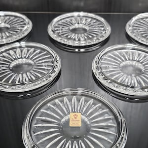 Op de afbeelding: Een set van zes ronde onderzetters van helder glas met een radiaal zonnestraalpatroon en gefacetteerde randen. Eén onderzetter heeft een klein vierkant goudkleurig label met de tekst Nachtmann en een kroonicoon in het midden.