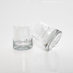 Peut inclure: Deux verres &agrave; whisky bas et transparents avec le logo "Johnnie Walker" imprim&eacute; en noir. Un verre est droit, l'autre est inclin&eacute;, tous deux sur une surface blanche.