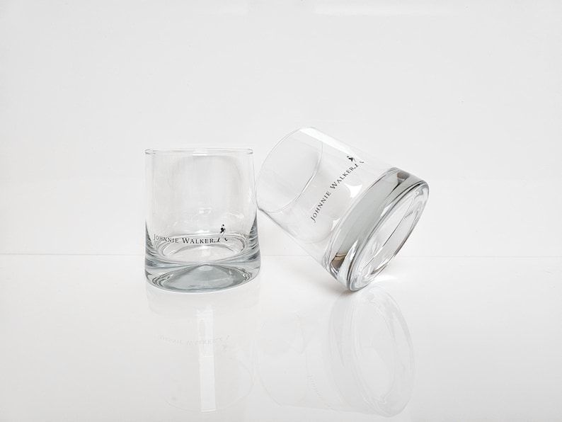 Peut inclure: Deux verres &agrave; whisky bas et transparents avec le logo "Johnnie Walker" imprim&eacute; en noir. Un verre est droit, l'autre est inclin&eacute;, tous deux sur une surface blanche.