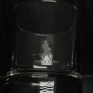 May include: Clear glass tumbler with the Johnnie Walker logo etched on the front. The logo features a silhouette of a man walking with a cane. The brand name is printed below the figure.