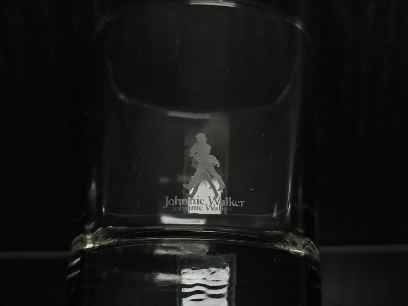 May include: Clear glass tumbler with the Johnnie Walker logo etched on the front. The logo features a silhouette of a man walking with a cane. The brand name is printed below the figure.