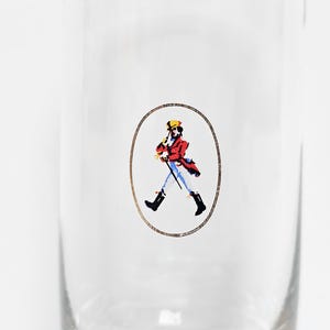 May include: Clear glass with a printed oval design featuring a figure in a red coat, yellow hat, and black boots, holding a cane. The figure is walking. The design is in a gold oval.