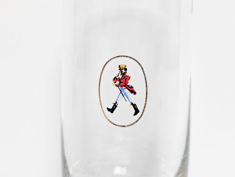 May include: Clear glass with a printed oval design featuring a figure in a red coat, yellow hat, and black boots, holding a cane. The figure is walking. The design is in a gold oval.