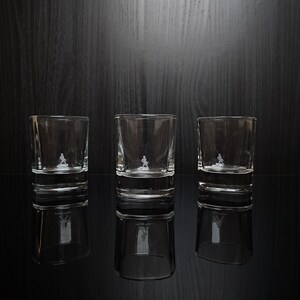 May include: Three clear, lowball glasses with a small, etched figure on the front. The glasses are reflected on a glossy black surface. The background is a dark wood grain.