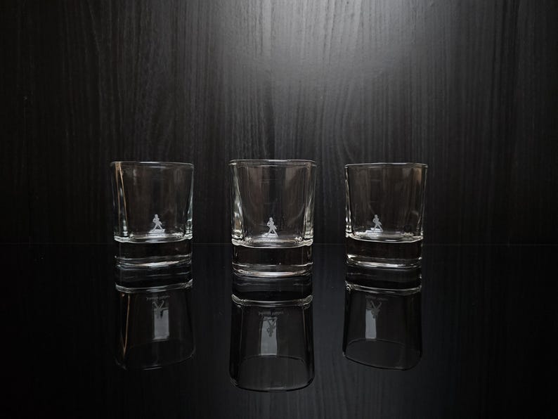 May include: Three clear, lowball glasses with a small, etched figure on the front. The glasses are reflected on a glossy black surface. The background is a dark wood grain.