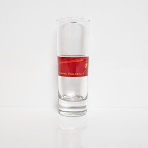 May include: A clear, tall shot glass with a red and gold band featuring the text "JOHNNIE WALKER." The glass is filled with a clear liquid, likely a spirit, and sits on a white surface. The design is simple and elegant.