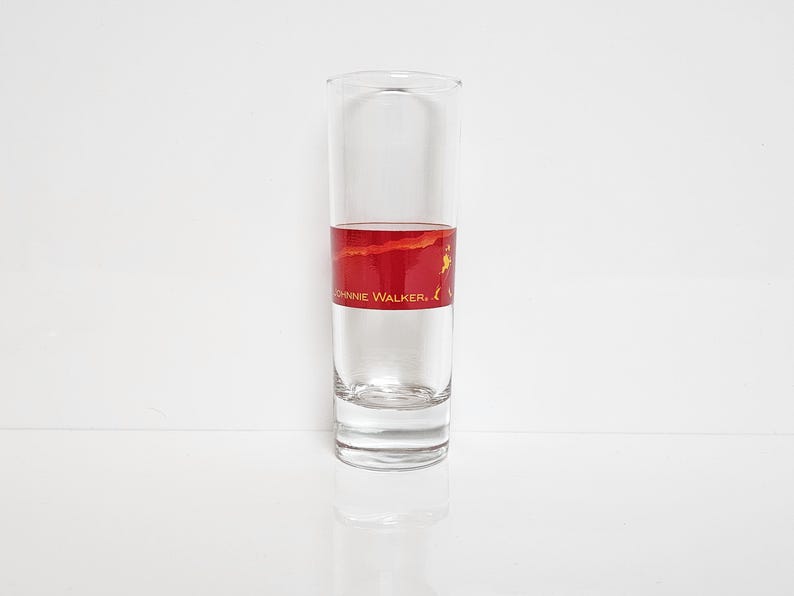 May include: A clear, tall shot glass with a red and gold band featuring the text "JOHNNIE WALKER." The glass is filled with a clear liquid, likely a spirit, and sits on a white surface. The design is simple and elegant.