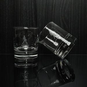 May include: Two clear, lowball whiskey glasses with etched designs. One glass is upright, the other tilted, and a third is reflected below. Each glass features a silhouette of a man walking with a top hat and the text "Johnnie Walker".