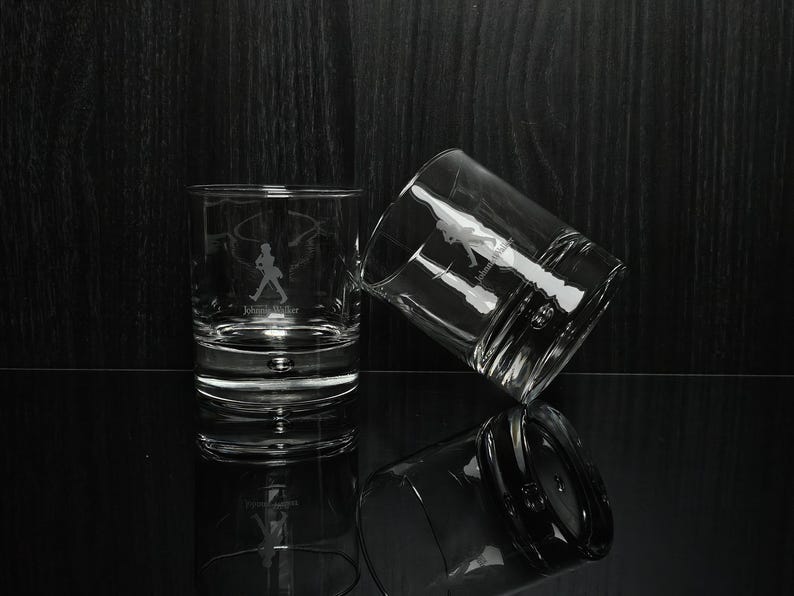 May include: Two clear, lowball whiskey glasses with etched designs. One glass is upright, the other tilted, and a third is reflected below. Each glass features a silhouette of a man walking with a top hat and the text "Johnnie Walker".