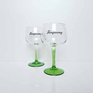 May include: Two clear, stemmed gin glasses with green stems and bases. The word "Tanqueray" is printed in black on the glass. The glasses are on a white surface.