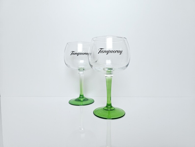 May include: Two clear, stemmed gin glasses with green stems and bases. The word "Tanqueray" is printed in black on the glass. The glasses are on a white surface.