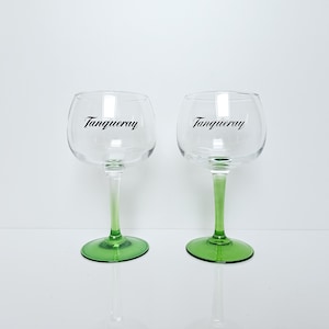 May include: Two clear, stemmed gin glasses with green bases and stems. The glasses have a rounded bowl shape and the word "Tanqueray" is printed in black on the front of each glass.