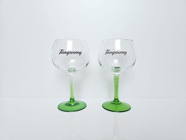 May include: Two clear, stemmed gin glasses with green bases and stems. The glasses have a rounded bowl shape and the word "Tanqueray" is printed in black on the front of each glass.