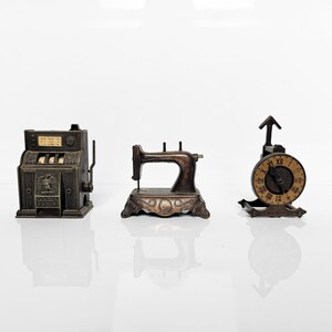 Pode incluir: Três miniaturas antigas de metal: uma caixa registradora com texto, uma máquina de costura e um relógio com algarismos romanos. Todos são castanhos e estão sobre uma superfície branca.