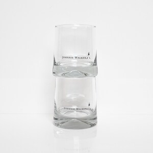 Peut inclure: Deux verres &agrave; boire cylindriques transparents empil&eacute;s l'un sur l'autre. Chaque verre porte l'inscription "JOHNNIE WALKER" en noir, ainsi qu'un petit logo d'un homme qui marche. Les verres sont pos&eacute;s sur une surface blanche.