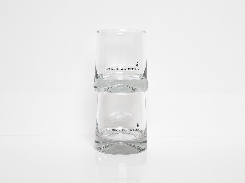 Peut inclure: Deux verres &agrave; boire cylindriques transparents empil&eacute;s l'un sur l'autre. Chaque verre porte l'inscription "JOHNNIE WALKER" en noir, ainsi qu'un petit logo d'un homme qui marche. Les verres sont pos&eacute;s sur une surface blanche.