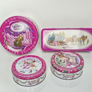 Op de afbeelding: Vier metalen Quality Street blikken in verschillende vormen en maten. Eén groot rond blik met het opschrift Year 2000 Special Edition, één rechthoekig blik met een afbeelding van een koets en twee kleinere ronde blikken met winterse taferelen.