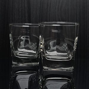 May include: Two clear square-shaped whiskey glasses with etched designs. Each glass features the words "KEEP WALKING" and a checkered flag design. The glasses are reflected on a dark surface.