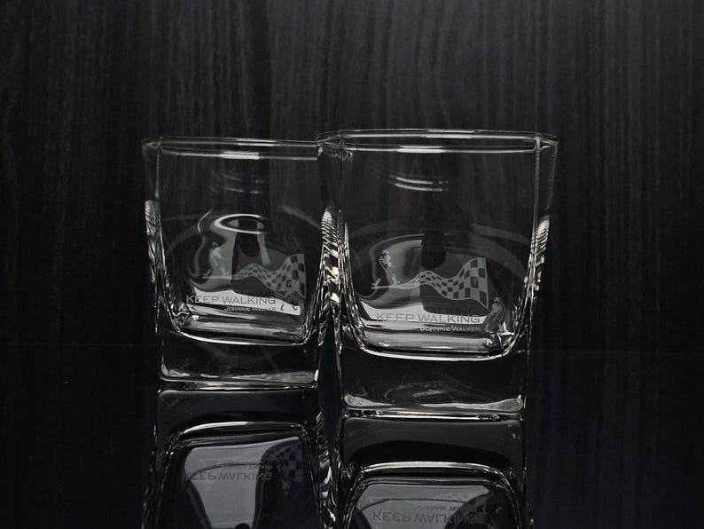 May include: Two clear square-shaped whiskey glasses with etched designs. Each glass features the words "KEEP WALKING" and a checkered flag design. The glasses are reflected on a dark surface.