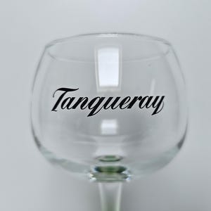 May include: A clear, stemmed gin glass with the word "Tanqueray" in black script across the bowl. The glass has a rounded bowl and a slender stem, set against a plain white background.