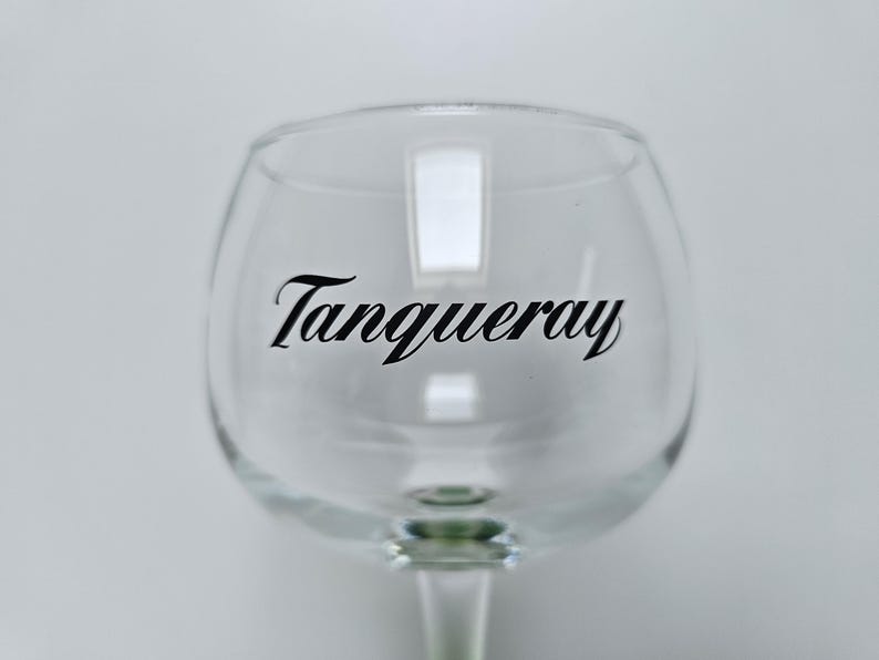 May include: A clear, stemmed gin glass with the word "Tanqueray" in black script across the bowl. The glass has a rounded bowl and a slender stem, set against a plain white background.