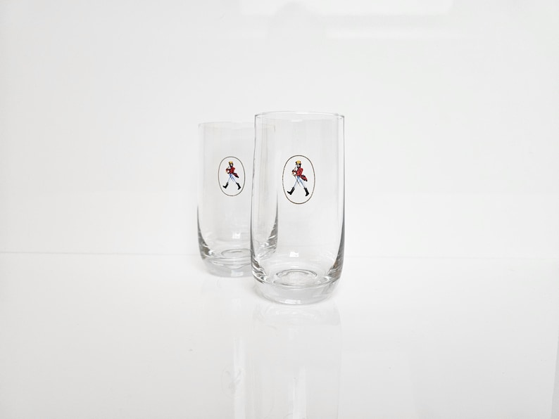 May include: Two clear, cylindrical drinking glasses. Each glass features a circular emblem with a figure in a red coat and black pants. The glasses are on a white surface.