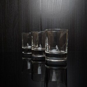 May include: Three clear shot glasses with a small etched figure of a person walking. The glasses are arranged in a row, with the largest glass in the foreground. The glasses are reflected on the black surface.
