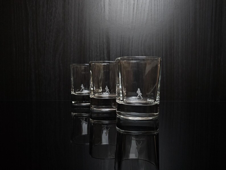May include: Three clear shot glasses with a small etched figure of a person walking. The glasses are arranged in a row, with the largest glass in the foreground. The glasses are reflected on the black surface.