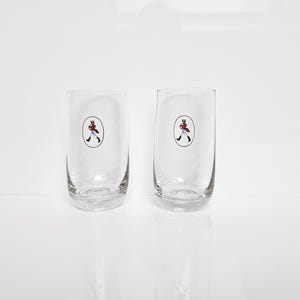 May include: Two clear, cylindrical drinking glasses. Each glass features a small, oval-shaped emblem with a figure in a red coat and black boots. The glasses are set against a white background, suggesting a clean, minimalist aesthetic.