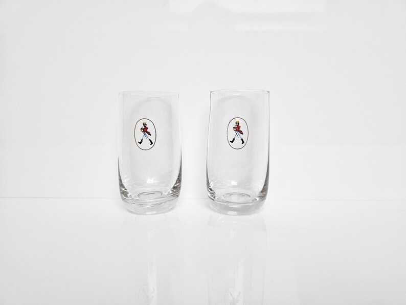 May include: Two clear, cylindrical drinking glasses. Each glass features a small, oval-shaped emblem with a figure in a red coat and black boots. The glasses are set against a white background, suggesting a clean, minimalist aesthetic.