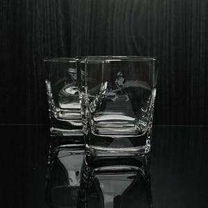 May include: Two clear, square-shaped whiskey glasses with etched designs. The glasses have a logo of a man walking with a cane. The glasses are on a reflective surface against a dark background.