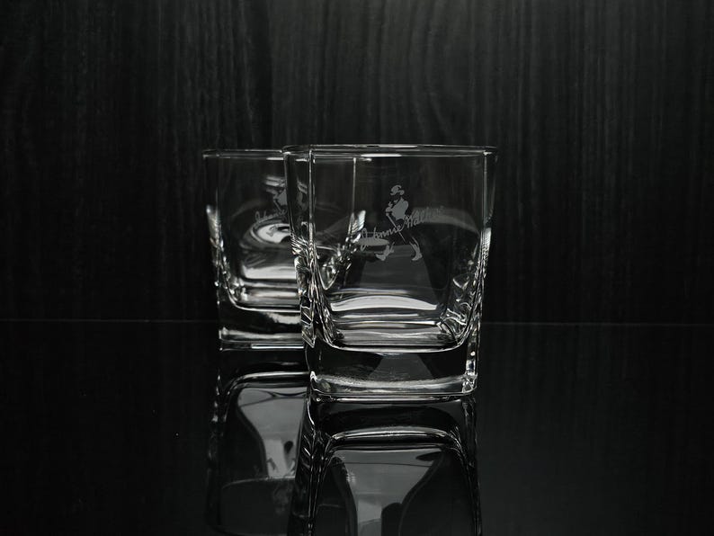 May include: Two clear, square-shaped whiskey glasses with etched designs. The glasses have a logo of a man walking with a cane. The glasses are on a reflective surface against a dark background.