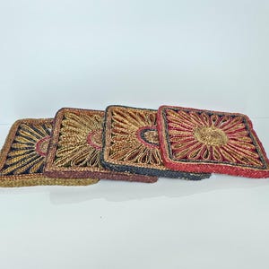 Peut inclure: Quatre dessous de verre carrés disposés en rangée, chacun présentant un motif floral tissé. Les dessous de verre affichent des combinaisons de couleurs variées, notamment or, bleu foncé, marron et rouge, avec un motif central circulaire.