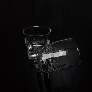 May include: Two clear, square-bottomed whiskey glasses on a black surface. One glass is upright, the other is tilted, with the text "KEEP WALKING" visible on the side. The glasses reflect on the glossy surface.