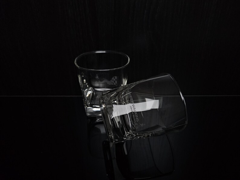 May include: Two clear, square-bottomed whiskey glasses on a black surface. One glass is upright, the other is tilted, with the text "KEEP WALKING" visible on the side. The glasses reflect on the glossy surface.