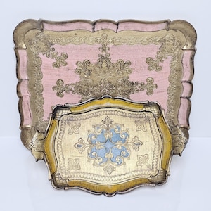 Op de afbeelding: Twee rechthoekige dienbladen met geschulpte randen. Het grotere dienblad heeft een roze middenstuk met gouden filigraanpatronen en een gouden rand. Het kleinere dienblad heeft een lichtblauw middenstuk met gouden bloemmotieven.