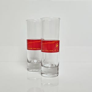 May include: Two clear, tall shot glasses with a red and gold band featuring the "Johnnie Walker" logo. The glasses are empty and stand on a white surface, reflecting the light.