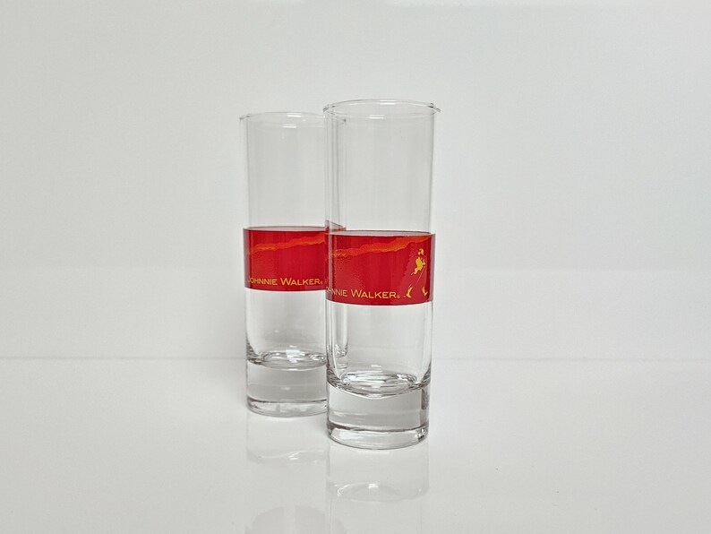 May include: Two clear, tall shot glasses with a red and gold band featuring the "Johnnie Walker" logo. The glasses are empty and stand on a white surface, reflecting the light.