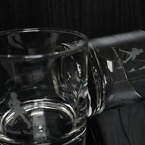 May include: Two clear, etched whiskey glasses with the "Johnnie Walker" logo and a silhouette of the Striding Man. The glasses are on a reflective surface, with one upright and the other tilted. The background is dark.