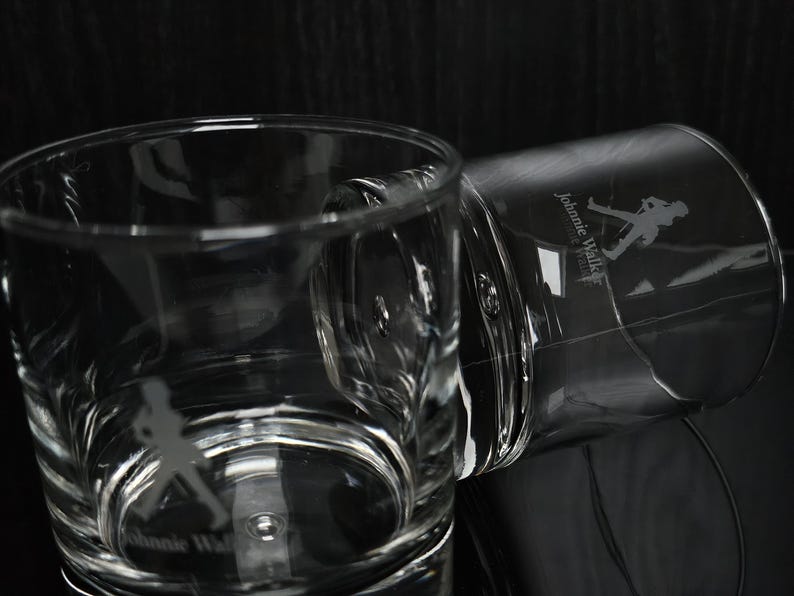 May include: Two clear, etched whiskey glasses with the "Johnnie Walker" logo and a silhouette of the Striding Man. The glasses are on a reflective surface, with one upright and the other tilted. The background is dark.