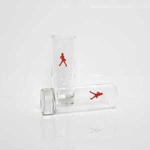 Peut inclure: Trois verres &agrave; shot transparents avec une figurine rouge d'un homme qui marche. Un verre est debout, un autre est couch&eacute; sur le c&ocirc;t&eacute;, et le troisi&egrave;me est la base d'un verre. Les verres sont sur une surface blanche.